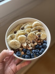 A hand holds a white bowl filled with a healthy assortment of sliced bananas, fresh blueberries, chia seeds, chocolate chips, chopped almonds, and walnuts.