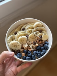 A hand holds a white bowl filled with a healthy assortment of sliced bananas, fresh blueberries, chia seeds, chocolate chips, chopped almonds, and walnuts.