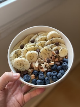 A hand holds a white bowl filled with a healthy assortment of sliced bananas, fresh blueberries, chia seeds, chocolate chips, chopped almonds, and walnuts.