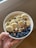 A hand holds a white bowl filled with a healthy assortment of sliced bananas, fresh blueberries, chia seeds, chocolate chips, chopped almonds, and walnuts.