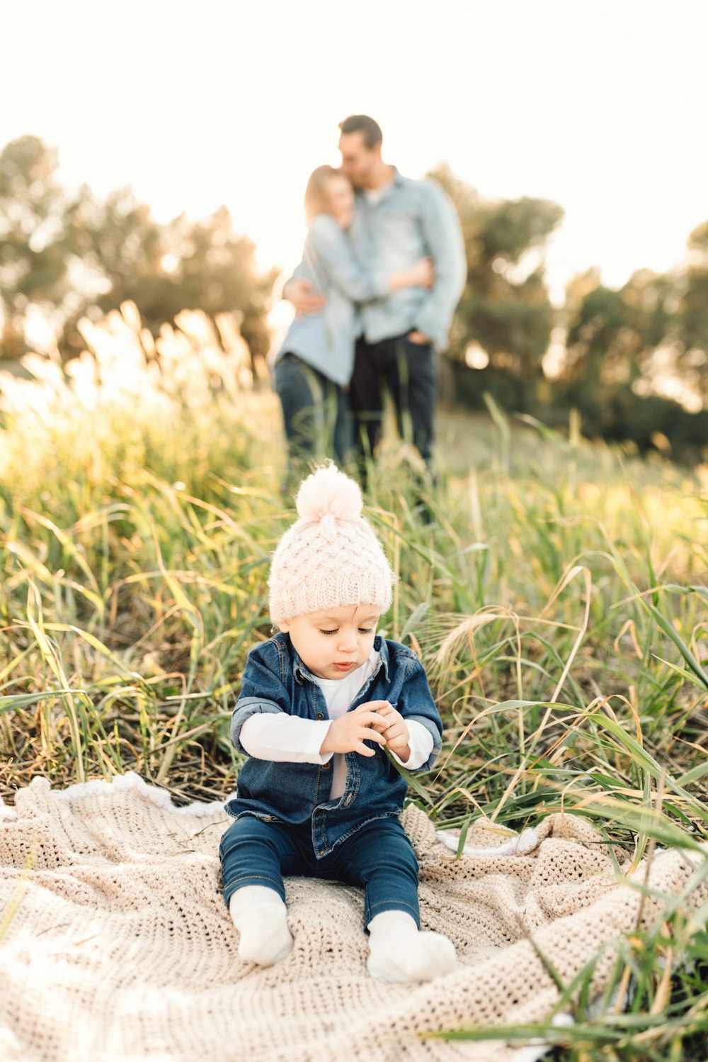 child in blue denim jacket and white knit cap sitting on white textile during daytime