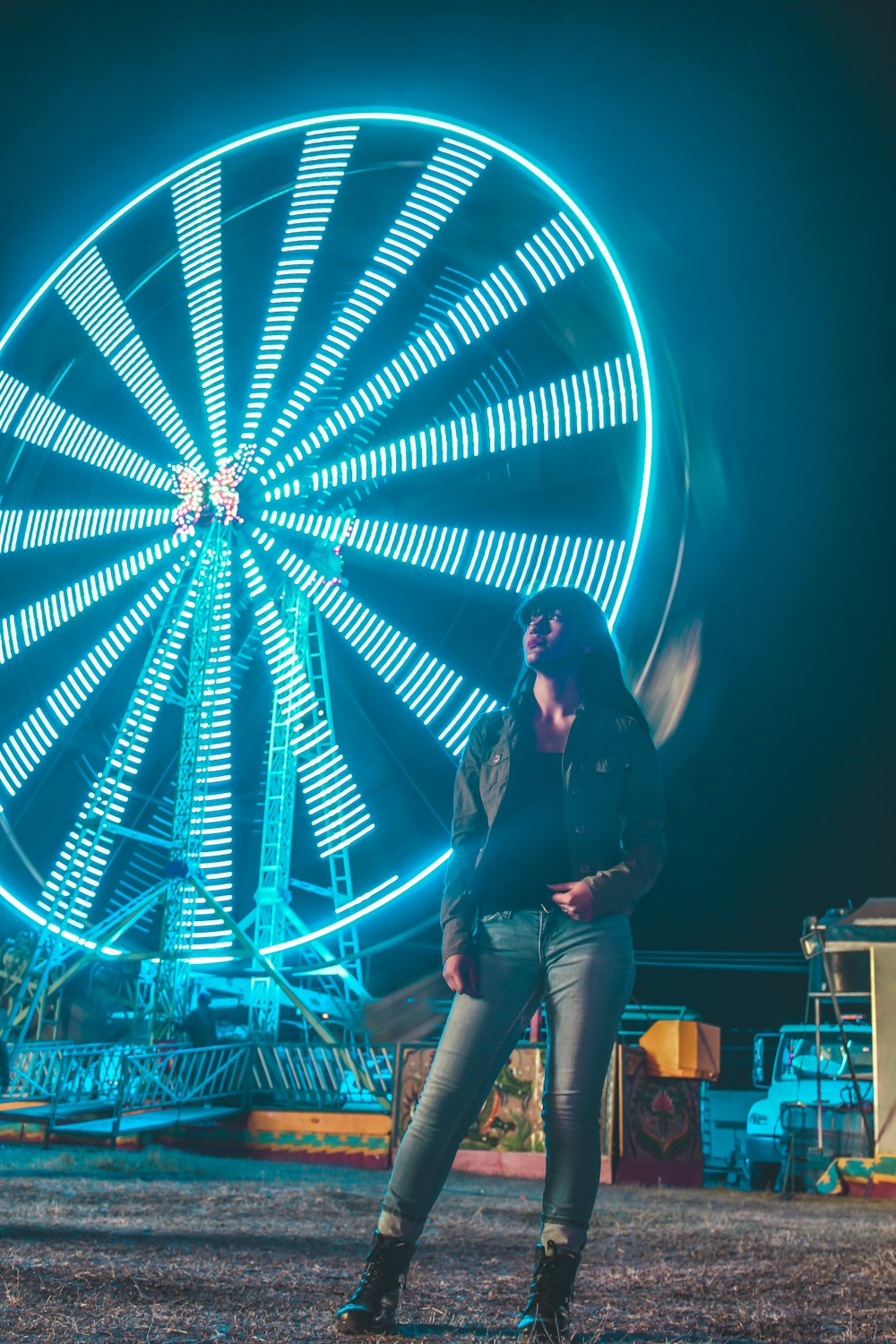 Mujer con chaqueta negra y jeans de mezclilla azul de pie cerca de la rueda de la fortuna iluminada azul durante la noche