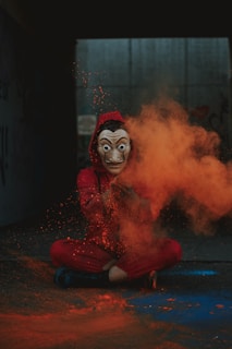 A person wearing a mask and a red hooded outfit sits cross-legged on the ground in a dark setting. Bright orange powder is explosively dispersed from their hands, creating a vivid contrast against the dim background.