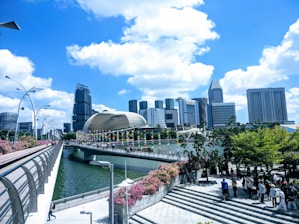 A modern cityscape featuring tall skyscrapers with unique architectural designs against a vibrant blue sky with white clouds. A bridge decorated with colorful banners crosses over a river, leading towards the urban buildings. Lush green trees and pink flowering bushes line a walkway where people are strolling.