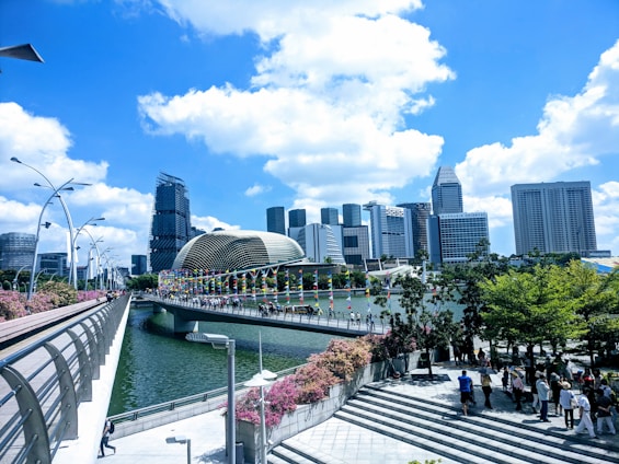 A modern cityscape featuring tall skyscrapers with unique architectural designs against a vibrant blue sky with white clouds. A bridge decorated with colorful banners crosses over a river, leading towards the urban buildings. Lush green trees and pink flowering bushes line a walkway where people are strolling.