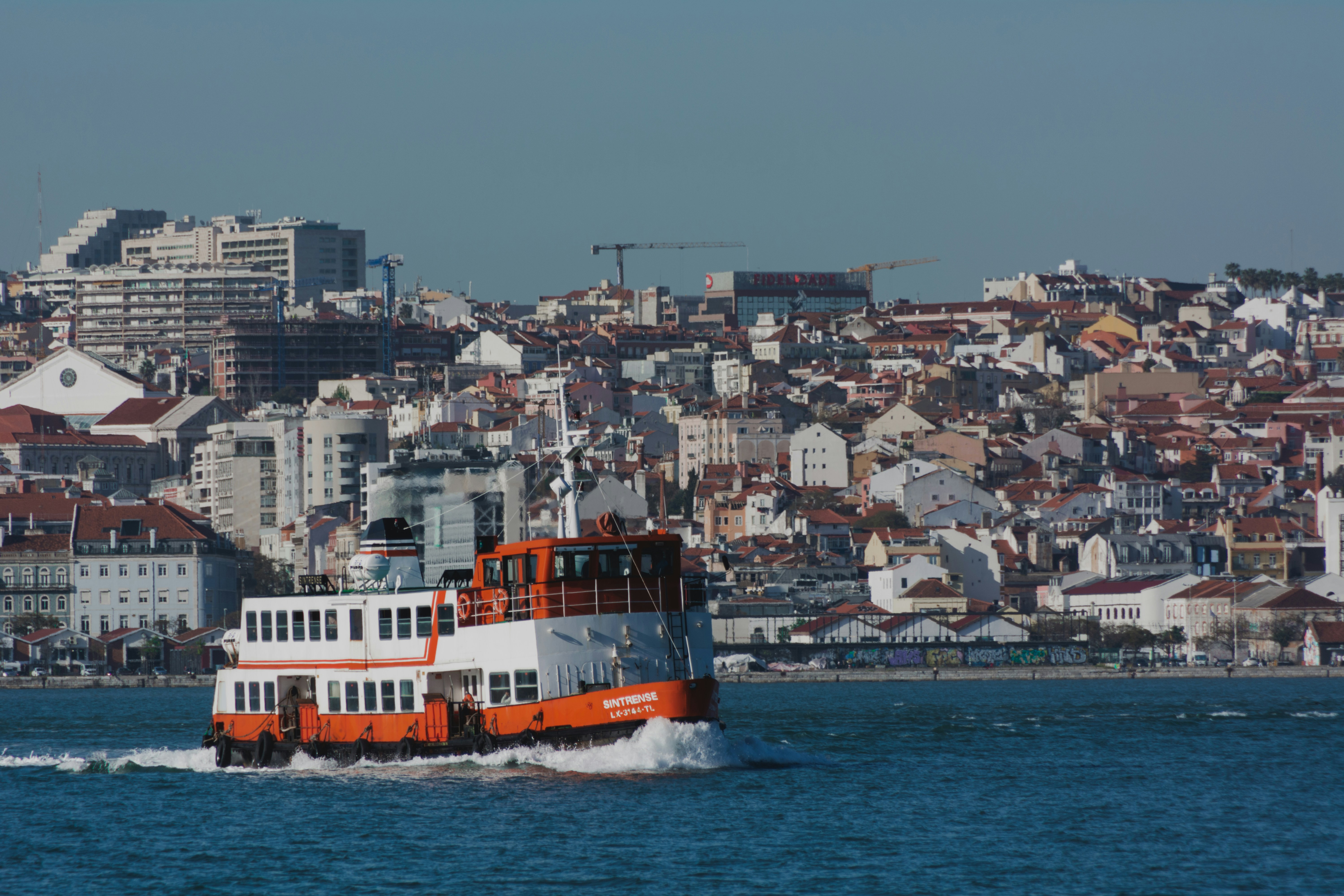 white and brown boat on sea during daytime, A public passenger ferry crossing the Tejo River in Lisbon, Portugal. The city is seen in the background.