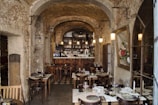 An interior of a rustic restaurant with stone walls and arched ceilings. The room is adorned with hanging pendant lights above a cozy bar, which is lined with a variety of bottles. Several wooden tables with neatly set plates, glasses, and cutlery are arranged throughout the space. A large framed floral painting hangs on one wall, adding a touch of elegance.