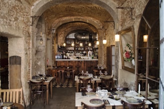 An interior of a rustic restaurant with stone walls and arched ceilings. The room is adorned with hanging pendant lights above a cozy bar, which is lined with a variety of bottles. Several wooden tables with neatly set plates, glasses, and cutlery are arranged throughout the space. A large framed floral painting hangs on one wall, adding a touch of elegance.