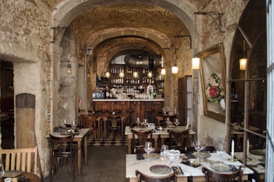 An interior of a rustic restaurant with stone walls and arched ceilings. The room is adorned with hanging pendant lights above a cozy bar, which is lined with a variety of bottles. Several wooden tables with neatly set plates, glasses, and cutlery are arranged throughout the space. A large framed floral painting hangs on one wall, adding a touch of elegance.