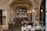 An interior of a rustic restaurant with stone walls and arched ceilings. The room is adorned with hanging pendant lights above a cozy bar, which is lined with a variety of bottles. Several wooden tables with neatly set plates, glasses, and cutlery are arranged throughout the space. A large framed floral painting hangs on one wall, adding a touch of elegance.