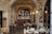 An interior of a rustic restaurant with stone walls and arched ceilings. The room is adorned with hanging pendant lights above a cozy bar, which is lined with a variety of bottles. Several wooden tables with neatly set plates, glasses, and cutlery are arranged throughout the space. A large framed floral painting hangs on one wall, adding a touch of elegance.