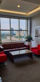 A well-lit office lounge area with large windows, offering a cityscape view. It features a brown leather sofa, two bright red armchairs, and a glass coffee table. Potted plants and framed artwork add to the decor.