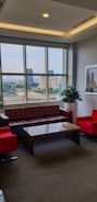 A well-lit office lounge area with large windows, offering a cityscape view. It features a brown leather sofa, two bright red armchairs, and a glass coffee table. Potted plants and framed artwork add to the decor.