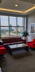 A well-lit office lounge area with large windows, offering a cityscape view. It features a brown leather sofa, two bright red armchairs, and a glass coffee table. Potted plants and framed artwork add to the decor.
