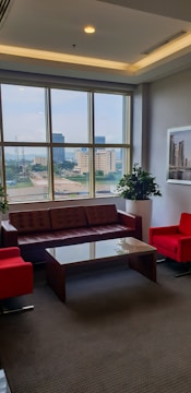 A well-lit office lounge area with large windows, offering a cityscape view. It features a brown leather sofa, two bright red armchairs, and a glass coffee table. Potted plants and framed artwork add to the decor.