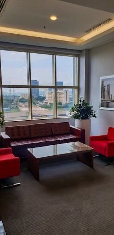 A well-lit office lounge area with large windows, offering a cityscape view. It features a brown leather sofa, two bright red armchairs, and a glass coffee table. Potted plants and framed artwork add to the decor.