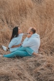 A couple sits playfully in a field of dry grass, facing each other and pulling on the strings of their hoodies. The scene conveys a sense of closeness and lighthearted interaction. Both are dressed in casual clothing with matching blue hoodies and jeans.