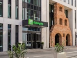 An entrance to a modern hotel building with a prominent brick wall feature. The hotel sign displays 'Holiday Inn' in green letters. Large glass windows are visible, and several potted plants decorate the area around the entrance. The building incorporates both contemporary and historical architectural elements.