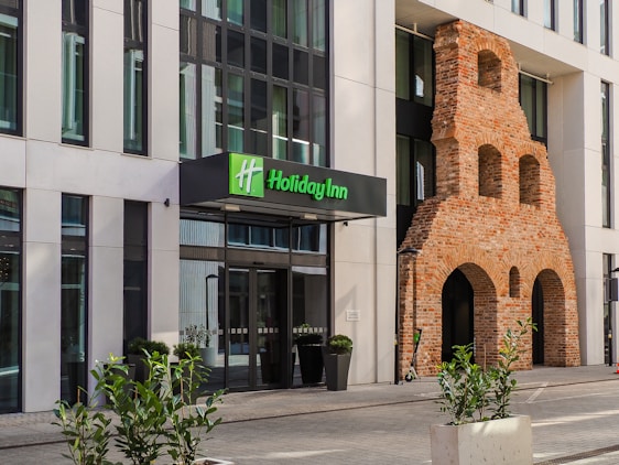 An entrance to a modern hotel building with a prominent brick wall feature. The hotel sign displays 'Holiday Inn' in green letters. Large glass windows are visible, and several potted plants decorate the area around the entrance. The building incorporates both contemporary and historical architectural elements.