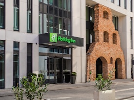 An entrance to a modern hotel building with a prominent brick wall feature. The hotel sign displays 'Holiday Inn' in green letters. Large glass windows are visible, and several potted plants decorate the area around the entrance. The building incorporates both contemporary and historical architectural elements.