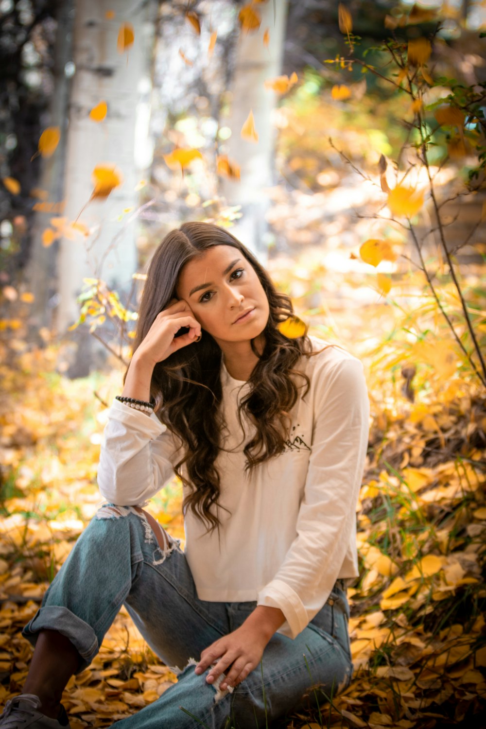 woman in white long sleeve shirt and blue denim jeans sitting on brown leaves during daytime