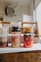 Several plastic containers filled with various colors of paint are stacked on a table. The background features a wooden crate and a softly lit interior space.