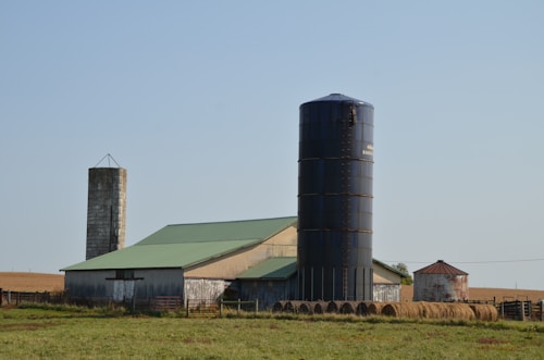 A rural farm setting featuring a large blue silo, a green-roofed barn, and a smaller metal silo. Hay bales are stacked in front of the structures, and the scene is set against a clear blue sky with a grassy field in the foreground.