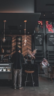 A dimly lit coffee shop interior features a person standing at the counter. The establishment has a modern design with wooden wall patterns and industrial-style lighting. A man wearing a traditional outfit is seated at a table in the background. There are decorative plants and posters with images of coffee beans on the walls.
