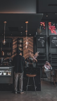A dimly lit coffee shop interior features a person standing at the counter. The establishment has a modern design with wooden wall patterns and industrial-style lighting. A man wearing a traditional outfit is seated at a table in the background. There are decorative plants and posters with images of coffee beans on the walls.