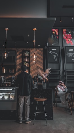 A dimly lit coffee shop interior features a person standing at the counter. The establishment has a modern design with wooden wall patterns and industrial-style lighting. A man wearing a traditional outfit is seated at a table in the background. There are decorative plants and posters with images of coffee beans on the walls.