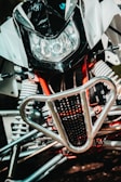 A close-up view of a motorbike's front section, featuring a large headlight, a black and white fairing, and a protective metallic grille. The intricate suspension springs and red framework parts are visible, showcasing mechanical details.