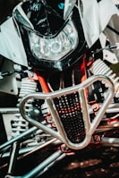 A close-up view of a motorbike's front section, featuring a large headlight, a black and white fairing, and a protective metallic grille. The intricate suspension springs and red framework parts are visible, showcasing mechanical details.