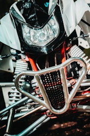 A close-up view of a motorbike's front section, featuring a large headlight, a black and white fairing, and a protective metallic grille. The intricate suspension springs and red framework parts are visible, showcasing mechanical details.