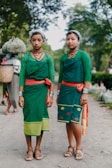 Two women wearing traditional green attire with red accents, adorned with necklaces and silver bracelets, stand on a dirt path surrounded by lush greenery. Their expressions are calm and they both have their hair tied back with a cloth. In the background, another person carries a basket on their back.