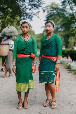 Two women wearing traditional green attire with red accents, adorned with necklaces and silver bracelets, stand on a dirt path surrounded by lush greenery. Their expressions are calm and they both have their hair tied back with a cloth. In the background, another person carries a basket on their back.