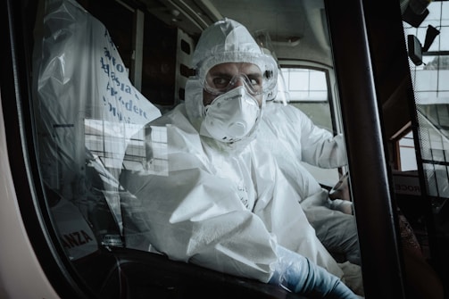 A person wearing full protective medical gear, including a hood and respirator mask, is seated inside what appears to be an ambulance. The interior is lit by outside light coming through the vehicle's windows.