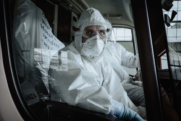 A person wearing full protective medical gear, including a hood and respirator mask, is seated inside what appears to be an ambulance. The interior is lit by outside light coming through the vehicle's windows.