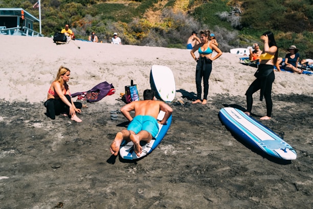 A beach scene with several people preparing for surfing. A man in blue shorts practices on a surfboard on the sand, while a woman in a red bikini top and black leggings sits nearby. Two other women, one in a blue bikini top and another in a yellow top, stand observing. Surfboards are laid out on the sand, and additional people relax on the dune in the background.