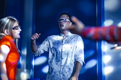 A group of people in a dimly lit environment, possibly a party or a club. The man in the center wears a patterned shirt and futuristic sunglasses. The woman to his left has long hair and wears a bright orange top, also with sunglasses. Soft, blue lighting and blurred figures in the background create a dynamic and energetic atmosphere. The presence of bright reflections and shadows suggests the use of vibrant, multicolored lights.