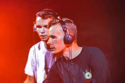 Two young individuals wearing headphones are focused on mixing music. They appear to be performing in a dimly lit environment with a strong red-orange lighting effect casting a vibrant glow around them.