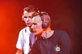 Two young individuals wearing headphones are focused on mixing music. They appear to be performing in a dimly lit environment with a strong red-orange lighting effect casting a vibrant glow around them.