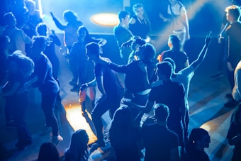 A group of people are dancing and socializing in a lively nightclub or party setting. The scene is illuminated by vibrant blue and orange lighting, creating a dynamic atmosphere. Some individuals have their arms raised, while others are engaging with one another, capturing a sense of enjoyment and celebration.