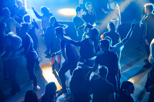 A group of people are dancing and socializing in a lively nightclub or party setting. The scene is illuminated by vibrant blue and orange lighting, creating a dynamic atmosphere. Some individuals have their arms raised, while others are engaging with one another, capturing a sense of enjoyment and celebration.