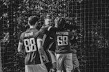 A group of soccer players are celebrating a goal, embracing each other near the goalpost. The image is in black and white, capturing the excitement and joy of the moment. The players wear jerseys with numbers and Cyrillic text on the back.