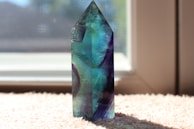 A polished, multicolored crystal with blue, green, and purple hues stands upright on a soft, textured surface near a window. Sunlight filters through the glass, casting light and slight shadows, highlighting the crystal's translucent and vibrant colors.