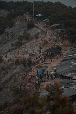 A hilly landscape features a winding pathway with people walking along it. Various makeshift buildings and tents are scattered alongside the path, surrounded by dense trees. Vendors with small stalls are set up, suggesting a local marketplace or tourist spot. The atmosphere appears overcast, lending a subdued tone to the scene.