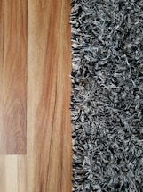 A close-up view showing the contrast between a smooth wooden floor and a textured, shaggy carpet. The left side features the warm tones and grain patterns of the wooden flooring, while the right side displays the irregular, intertwined fibers of a multi-toned black and gray carpet.