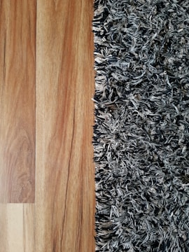 A close-up view showing the contrast between a smooth wooden floor and a textured, shaggy carpet. The left side features the warm tones and grain patterns of the wooden flooring, while the right side displays the irregular, intertwined fibers of a multi-toned black and gray carpet.