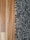A close-up view showing the contrast between a smooth wooden floor and a textured, shaggy carpet. The left side features the warm tones and grain patterns of the wooden flooring, while the right side displays the irregular, intertwined fibers of a multi-toned black and gray carpet.