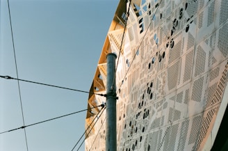 The image displays an intricate architectural facade with metallic panels featuring various perforated designs and patterns. A metallic pole intersects the view, with multiple cables connected to it, set against a clear blue sky.